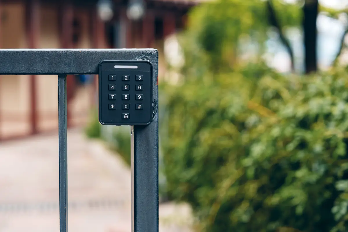 Clavier à code noir fixé sur un poteau à l’entrée d’un jardin, portail en arrière-plan